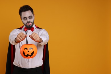 Photo of Man in scary vampire costume with fangs and pumpkin bucket on orange background, space for text. Halloween celebration