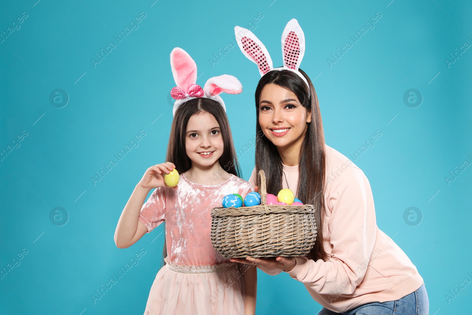 Photo of Mother and daughter in bunny ears headbands with basket of Easter eggs on color background