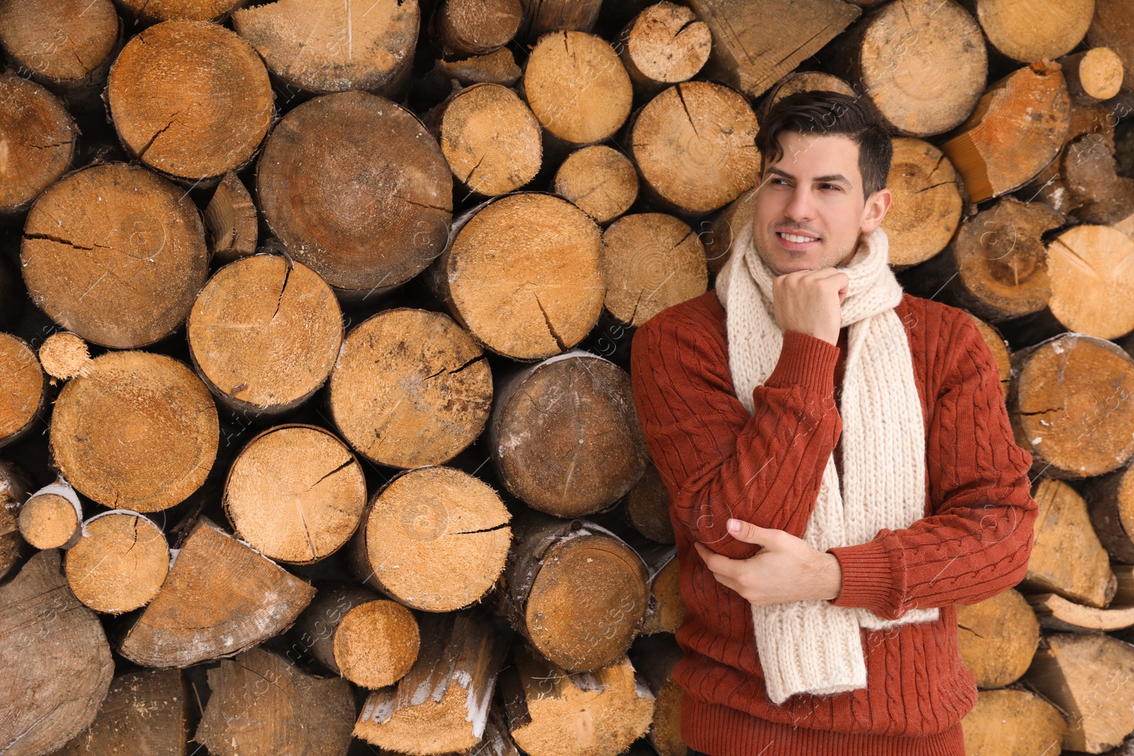 Photo of Handsome man wearing warm sweater and scarf near stack of firewood. Winter season