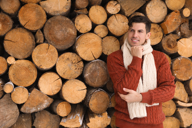 Photo of Handsome man wearing warm sweater and scarf near stack of firewood. Winter season