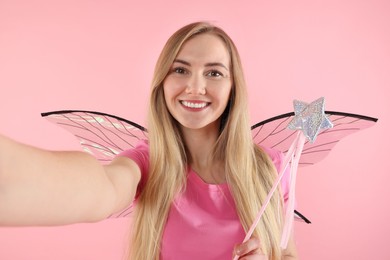 Photo of Beautiful girl in fairy costume with wings and magic wand taking selfie on pink background