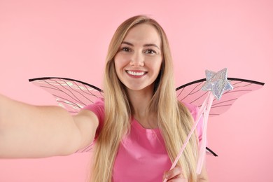 Beautiful girl in fairy costume with wings and magic wand taking selfie on pink background