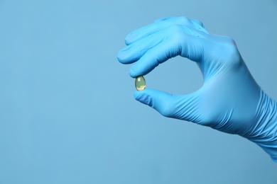 Scientist in protective gloves holding pill on light blue background, closeup. Space for text