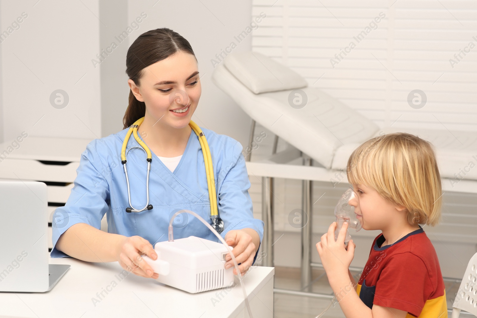 Photo of Medical assistant sitting near sick little boy while he using nebulizer for inhalation in hospital