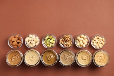 Photo of Many tasty nut butters in jars and nuts on brown table, flat lay. Space for text