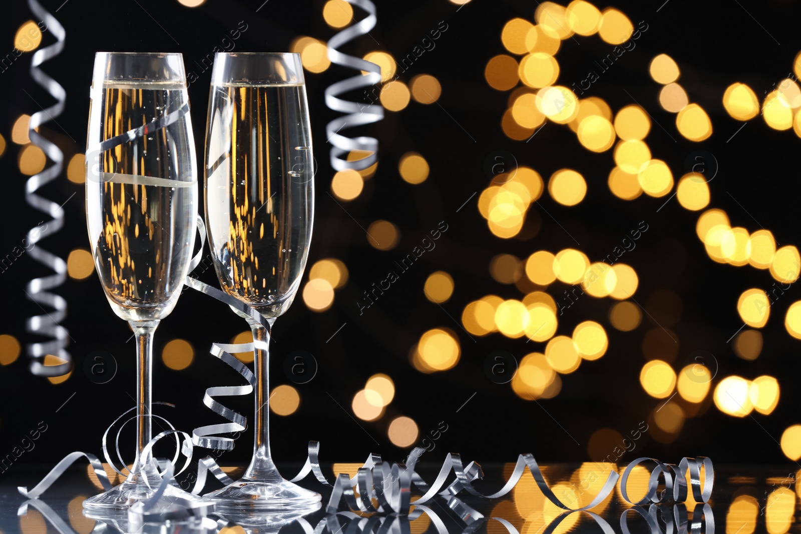 Photo of Glasses of champagne and serpentine streamers against black background with blurred lights. Space for text