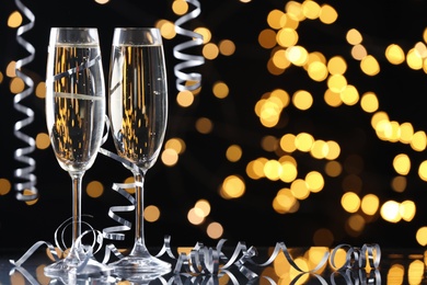Glasses of champagne and serpentine streamers against black background with blurred lights. Space for text
