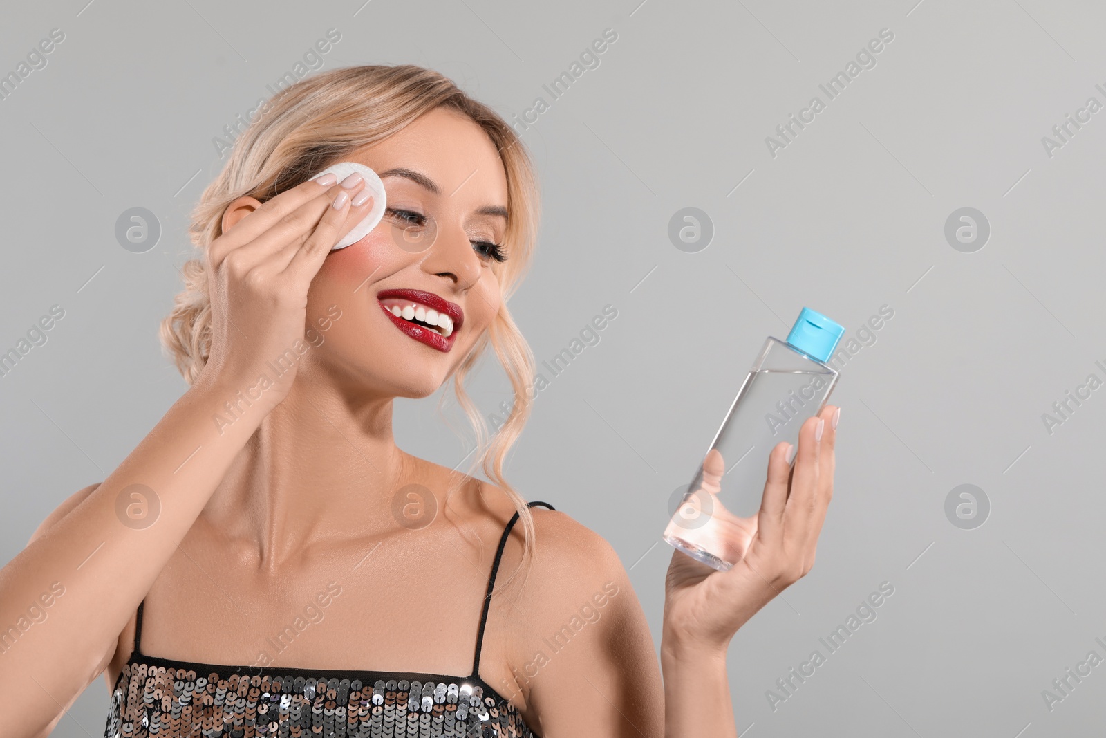 Photo of Smiling woman removing makeup with cotton pad and holding bottle on light grey background