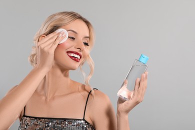 Photo of Smiling woman removing makeup with cotton pad and holding bottle on light grey background
