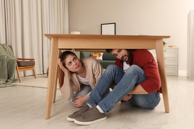 Scared couple hiding under table in living room during earthquake