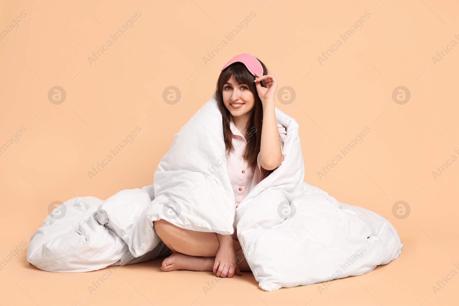 Photo of Happy woman with pyjama and sleep mask wrapped in blanket on beige background