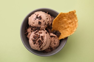 Tasty chocolate ice cream and piece of waffle cone in bowl on light green background, top view