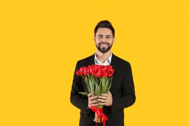 Photo of Happy man holding red tulip bouquet on yellow background. 8th of March celebration