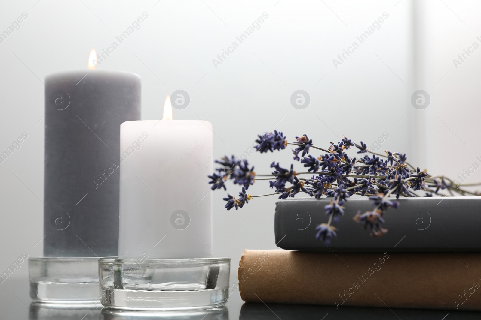 Photo of Wax candles in glass holders near books and lavender flowers on table against light background