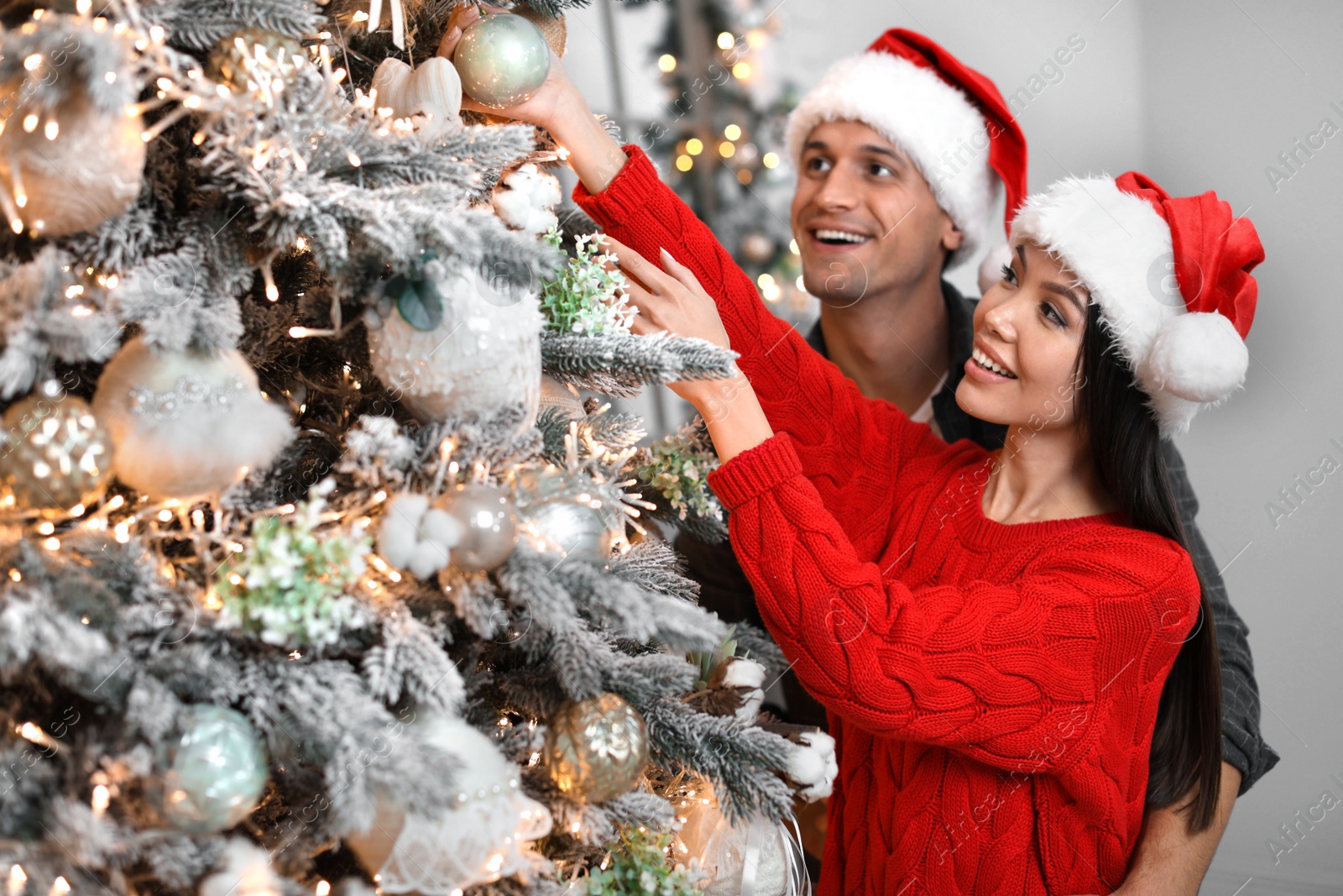 Photo of Happy young couple in Santa hats decorating Christmas tree together at home