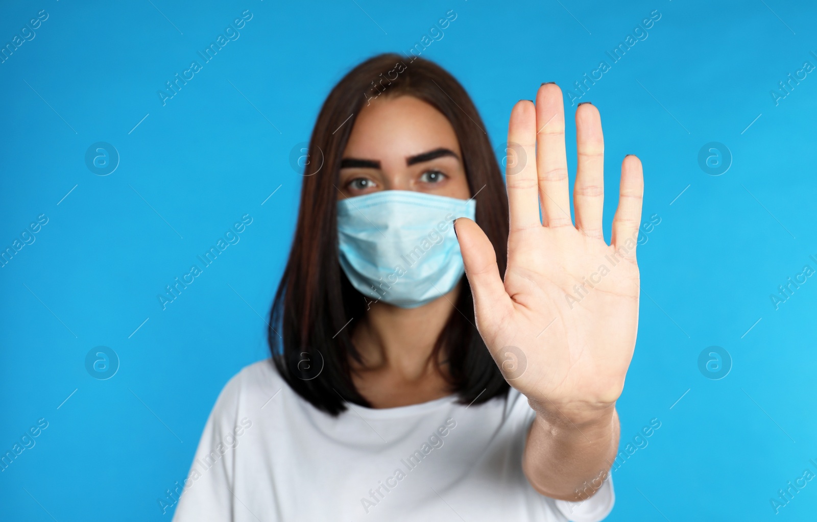 Photo of Young woman in protective mask showing stop gesture on light blue background. Prevent spreading of coronavirus