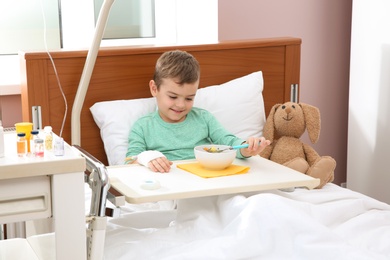 Photo of Little child with intravenous drip eating soup in hospital bed