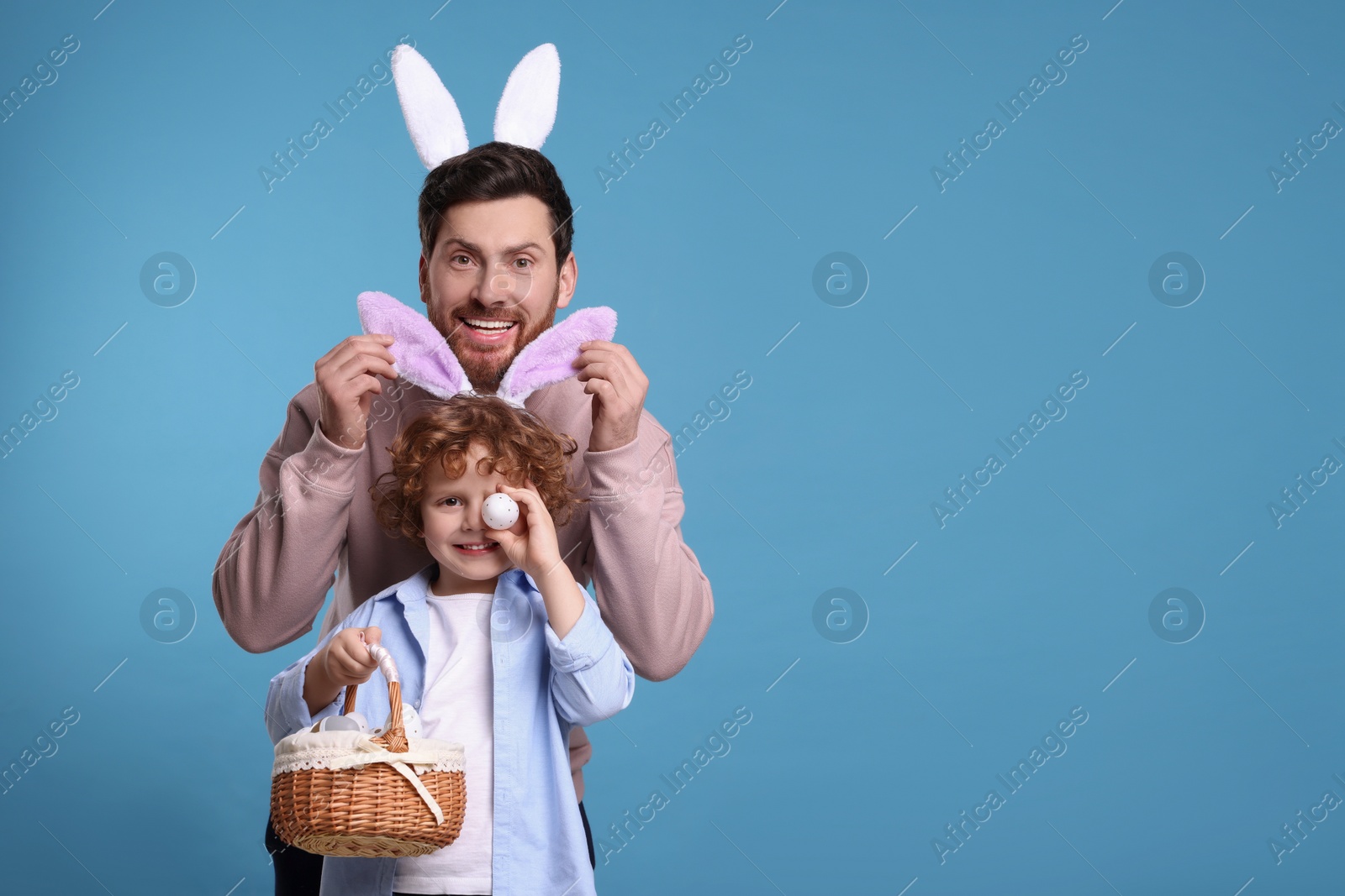 Photo of Happy father and son in cute bunny ears having fun on light blue background. Space for text. Easter celebration