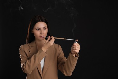 Woman using long cigarette holder for smoking on black background, space for text