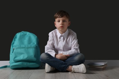 Photo of Little boy with backpack sitting on floor near black wall. School bullying