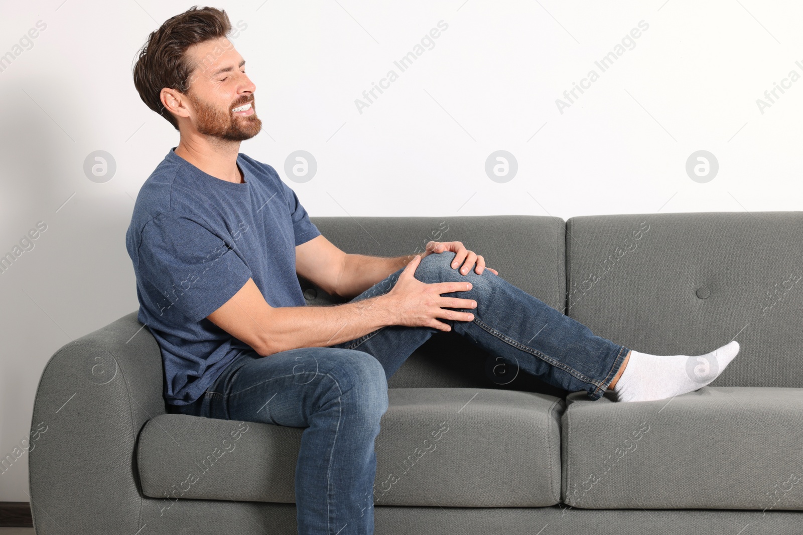 Photo of Man suffering from leg pain and touching knee on sofa indoors