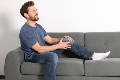 Man suffering from leg pain and touching knee on sofa indoors