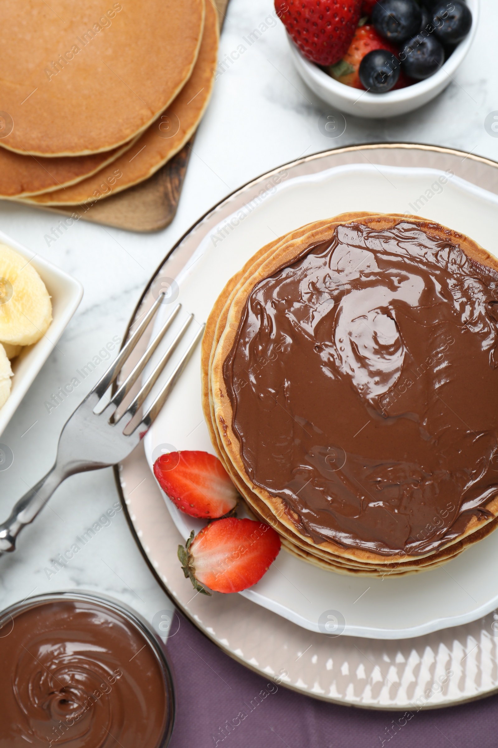 Photo of Tasty pancakes with chocolate paste, berries and fork on white marble table, flat lay