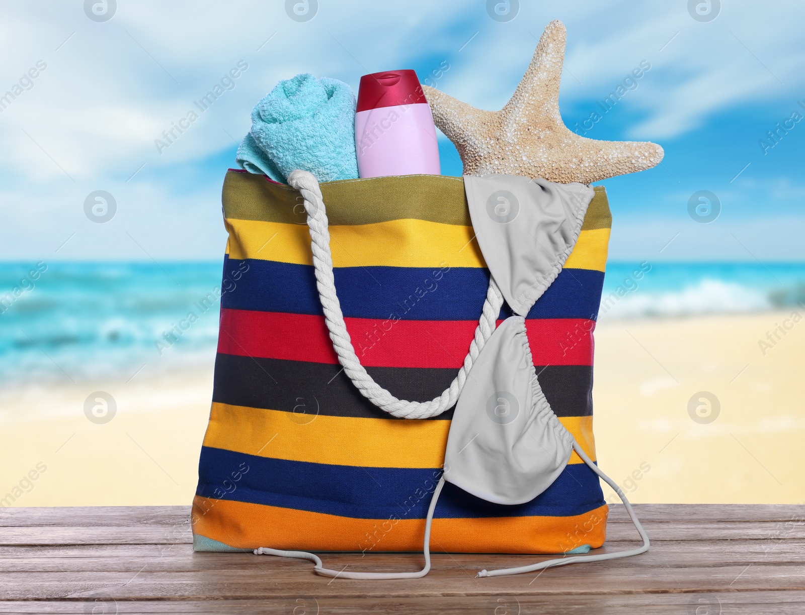 Image of Stylish striped bag and other beach accessories on wooden table near sea