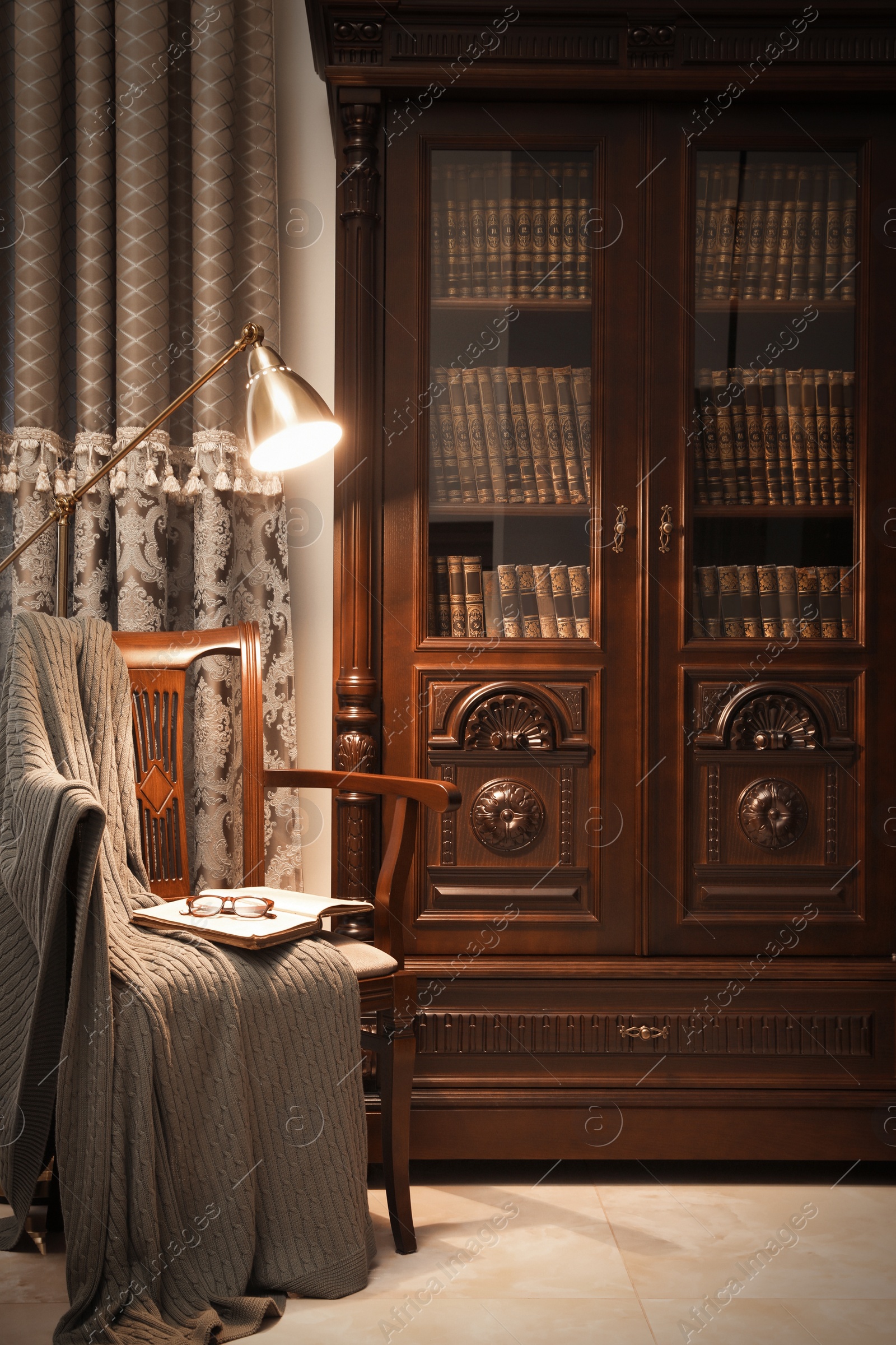 Photo of Comfortable armchair with book, blanket and lamp near wooden bookcase in library