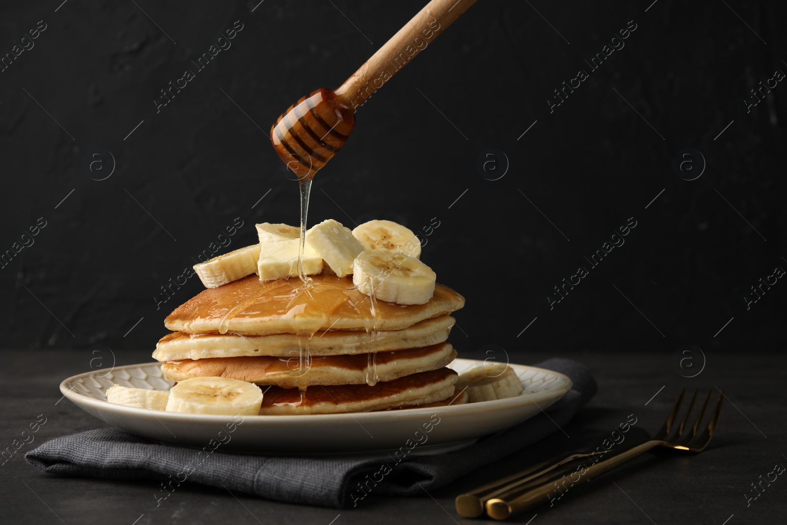 Photo of Pouring honey from dipper onto delicious pancakes with bananas and butter at dark table, space for text