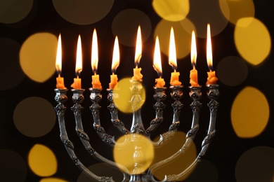 Photo of Silver menorah with burning candles against dark background and blurred festive lights. Hanukkah celebration