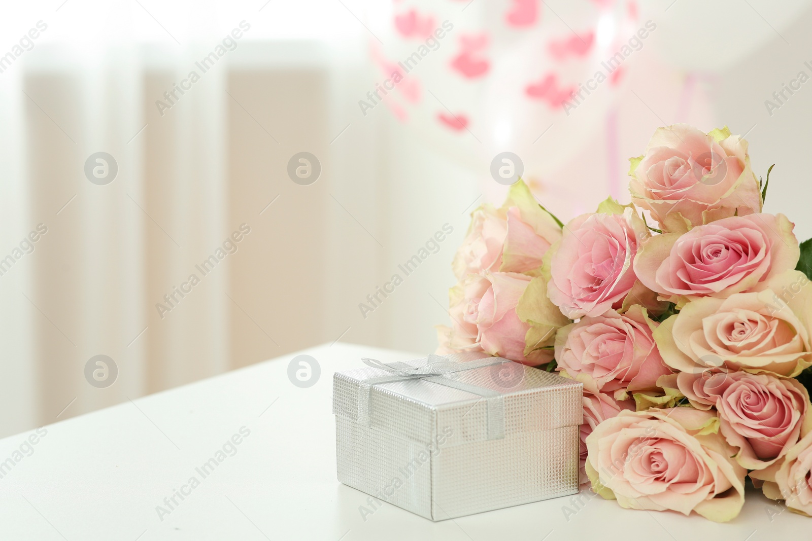 Photo of Beautiful bouquet of roses and gift on white table in room, space for text. Happy birthday greetings