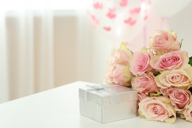 Photo of Beautiful bouquet of roses and gift on white table in room, space for text. Happy birthday greetings