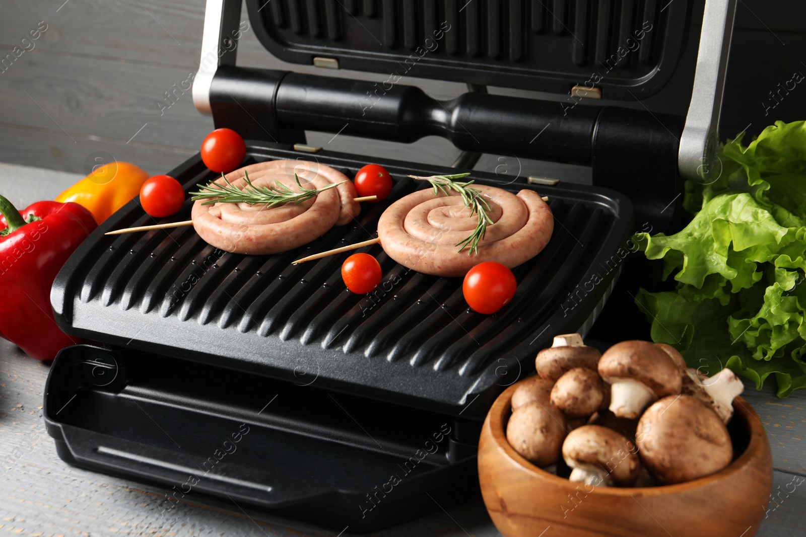 Photo of Electric grill with homemade sausages, rosemary and vegetables on rustic wooden table