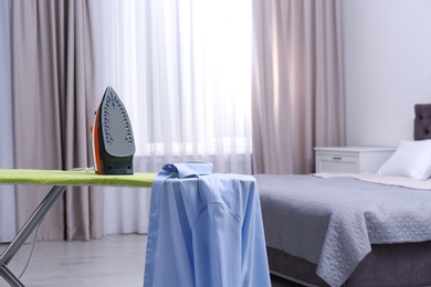 Photo of Modern electric iron and shirt on board in bedroom, space for text. Laundry day