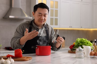 Photo of Man with pot smelling dish after cooking at countertop in kitchen