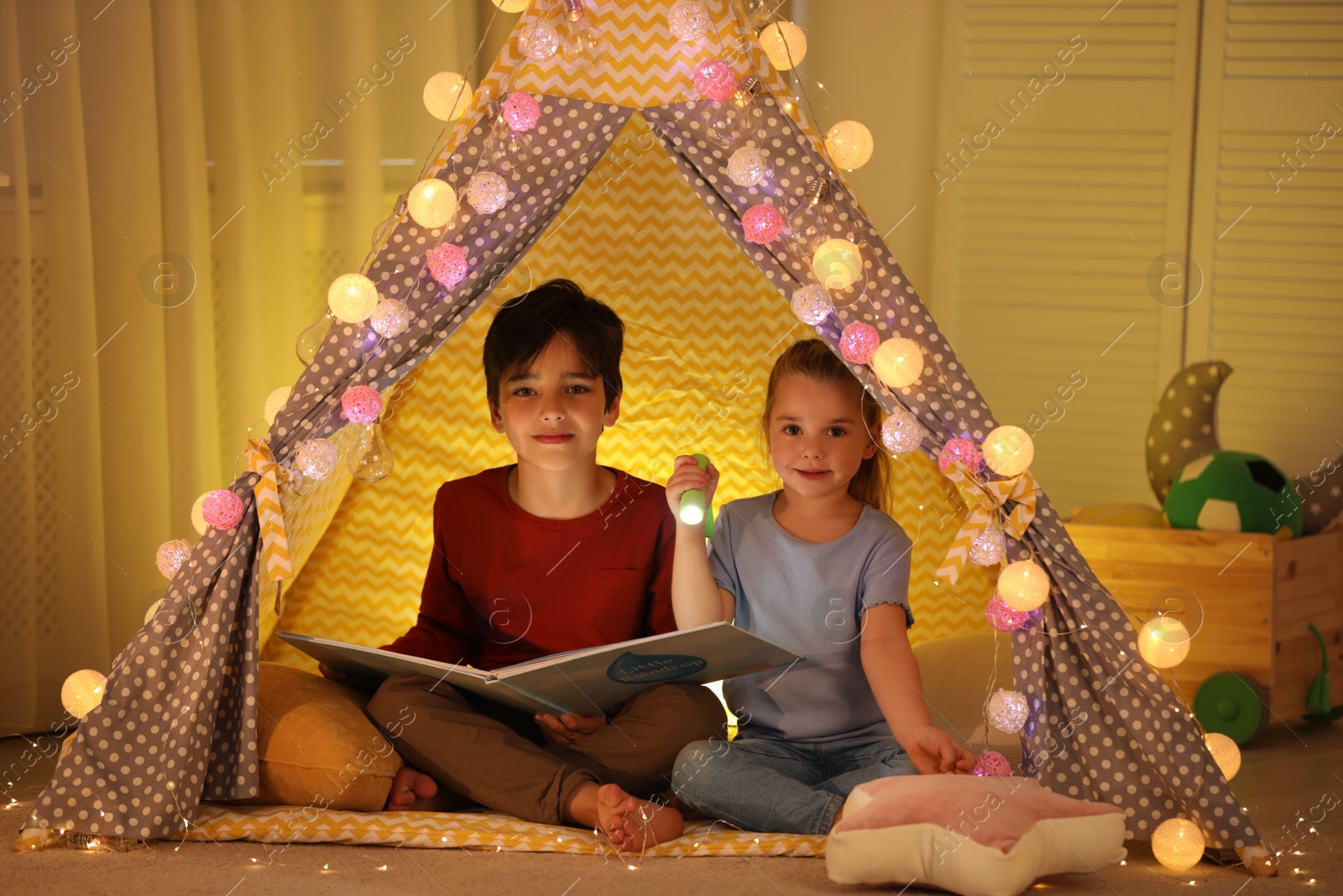 Photo of Little children with flashlight reading book in play tent at home