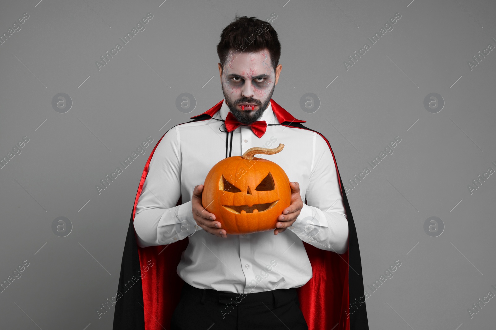 Photo of Man in scary vampire costume with fangs and carved pumpkin on light grey background. Halloween celebration