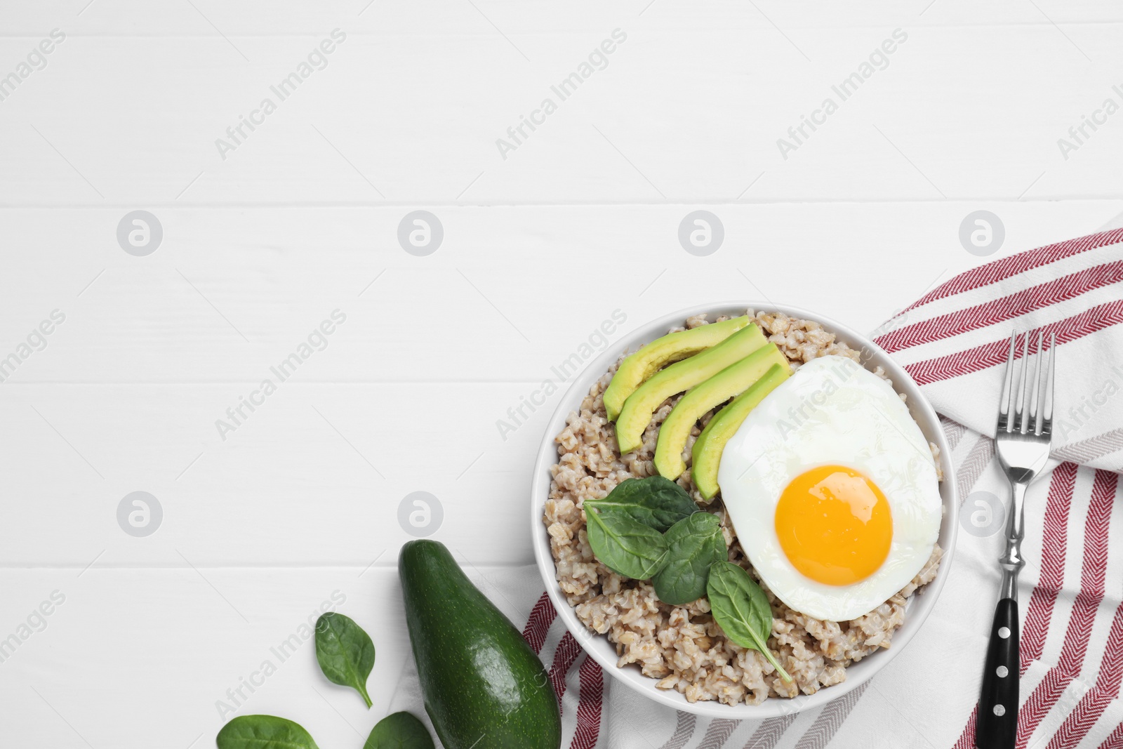 Photo of Tasty boiled oatmeal with fried egg, avocado and basil served on white wooden table, flat lay. Space for text