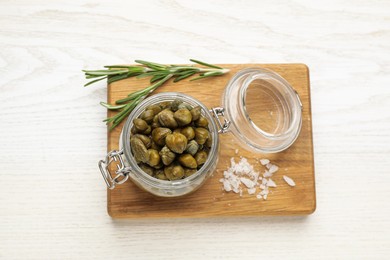 Jar with tasty capers, salt and rosemary on white wooden table, top view