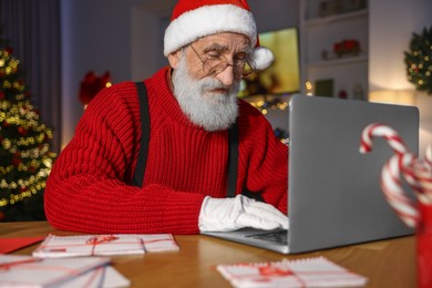 Photo of Santa Claus using laptop at his workplace in room decorated for Christmas