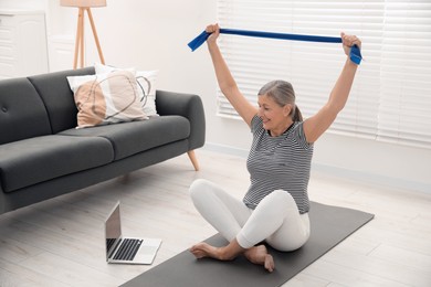 Senior woman doing exercise with fitness elastic band near laptop on mat at home