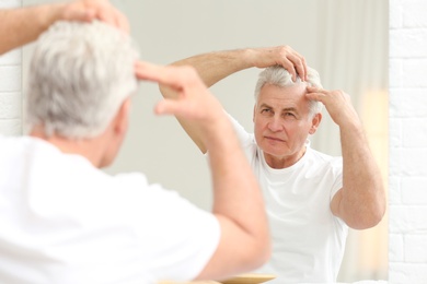 Photo of Senior man with hair loss problem looking in mirror indoors