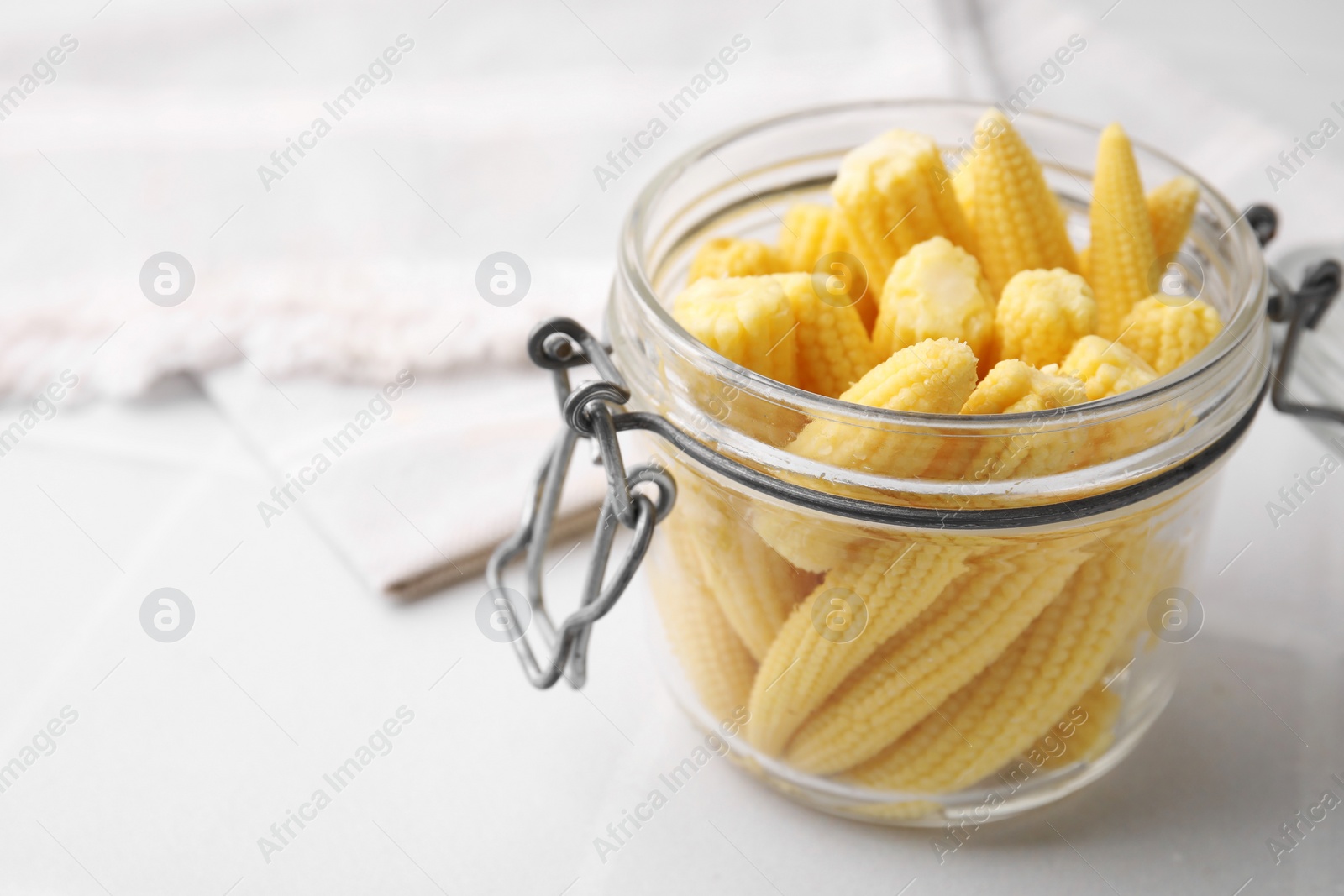 Photo of Tasty fresh yellow baby corns in glass jar on white tiled table, closeup. Space for text