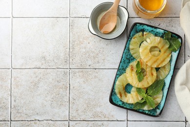 Tasty grilled pineapple slices, honey and wooden spoon on light gray tiled table, flat lay. Space for text