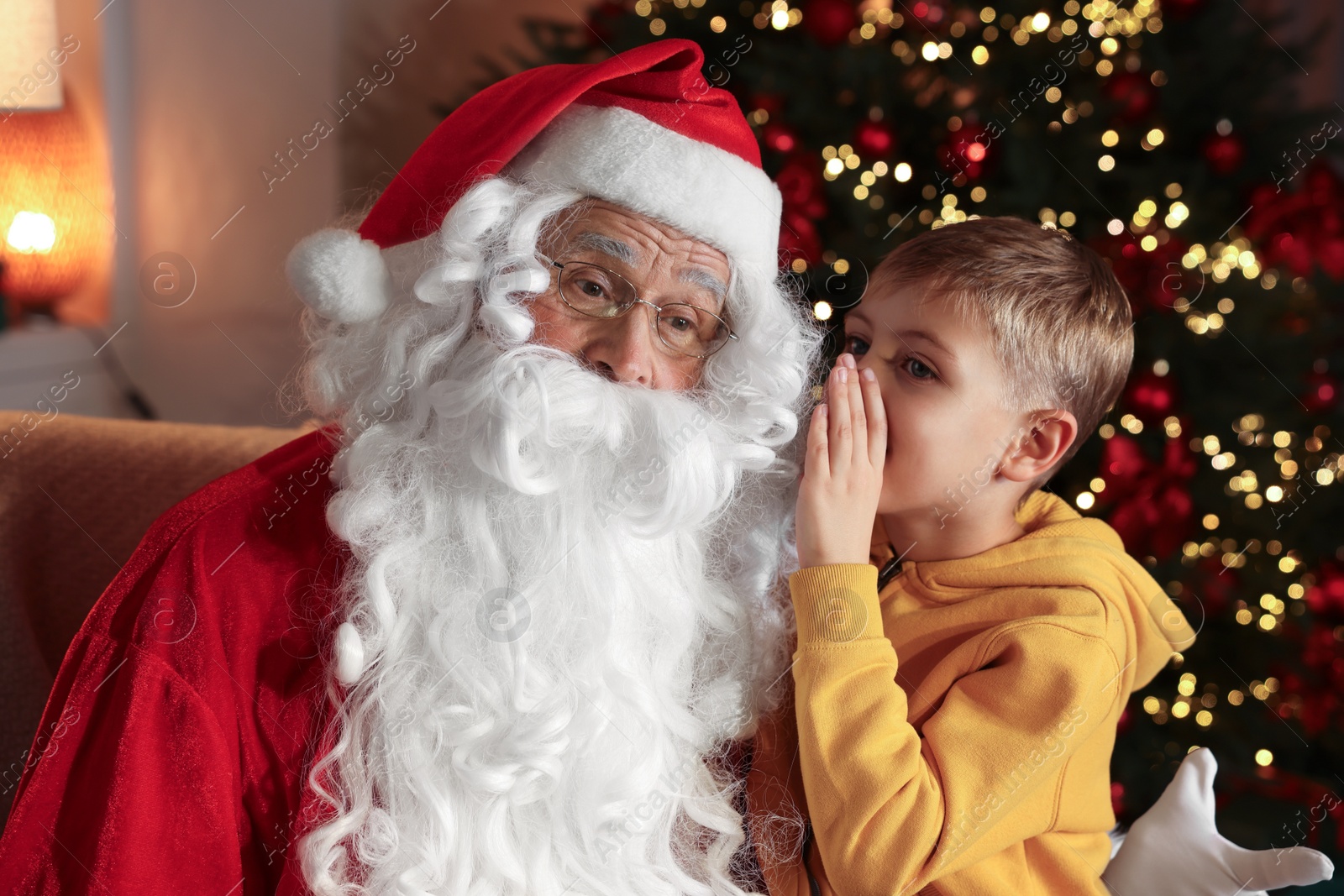 Photo of Little boy whispering his wish to Santa near Christmas tree in room