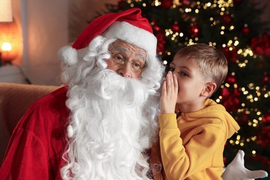 Photo of Little boy whispering his wish to Santa near Christmas tree in room