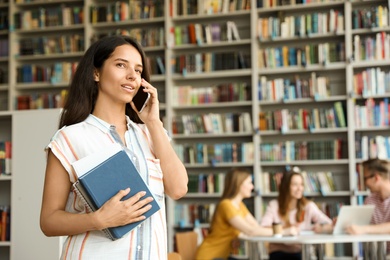 Photo of Young woman with books talking on phone in library. Space for text