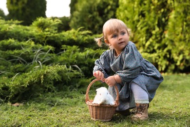 Cute little girl holding wicker basket with adorable rabbit outdoors on sunny day