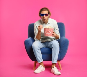 Photo of Emotional man with 3D glasses and popcorn sitting in armchair during cinema show on color background